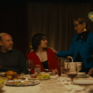 A group of people gathers around a dinner table, their attention fixed on a young woman seated at its head. She sits in the spotlight of the moment, while an older woman stands beside her, resting a hand gently on her shoulder. Seated next to her is a man wearing a yarmulke.