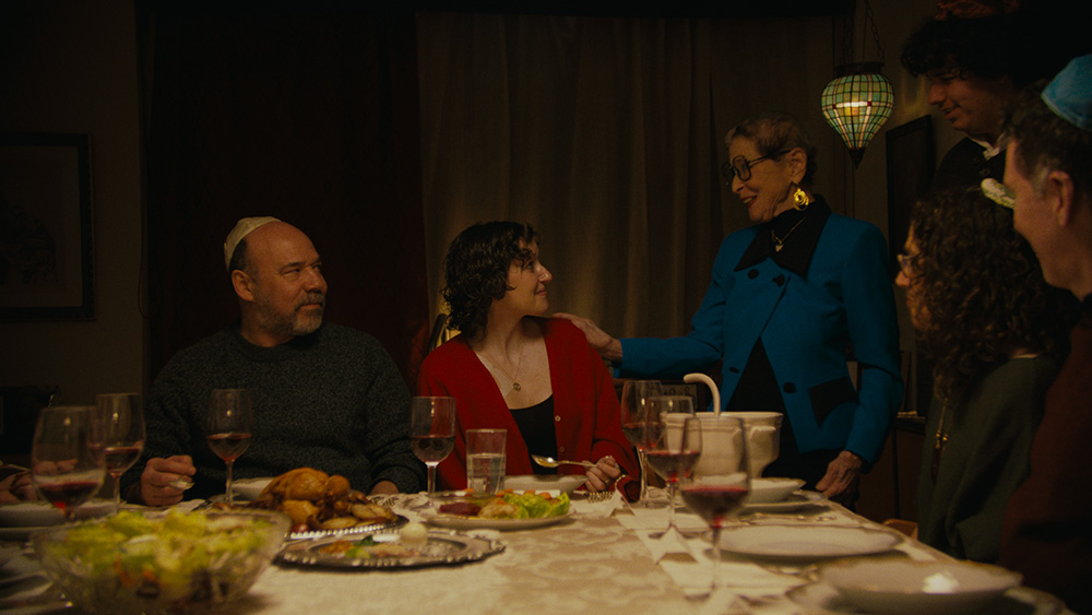 A group of people gathers around a dinner table, their attention fixed on a young woman seated at its head. She sits in the spotlight of the moment, while an older woman stands beside her, resting a hand gently on her shoulder. Seated next to her is a man wearing a yarmulke.