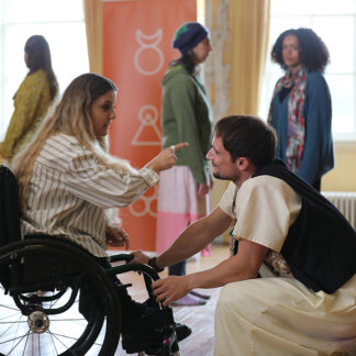 A group of people in a room with wooden floors and large windows with yellow curtains. One woman is sitting in a wheelchair, wearing a striped shirt, and is pointing at a man who is kneeling in front of the wheelchair. The kneeling person is wearing a white outfit with a black vest. Theother people n the background are standing and talking.