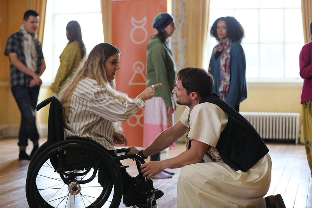 A group of people in a room with wooden floors and large windows with yellow curtains. One woman is sitting in a wheelchair, wearing a striped shirt, and is pointing at a man who is kneeling in front of the wheelchair. The kneeling person is wearing a white outfit with a black vest. Theother people n the background are standing and talking.
