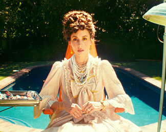 A woman with an expression of shock or surprise, wearing vintage-looking pearl necklaces, and gold earrings, and has an elaborate curly updo. There is a bright outdoor setting, with a pool and greenery in the background.