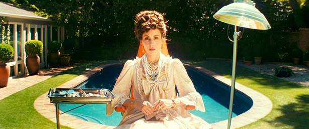 A woman with an expression of shock or surprise, wearing vintage-looking pearl necklaces, and gold earrings, and has an elaborate curly updo. There is a bright outdoor setting, with a pool and greenery in the background.