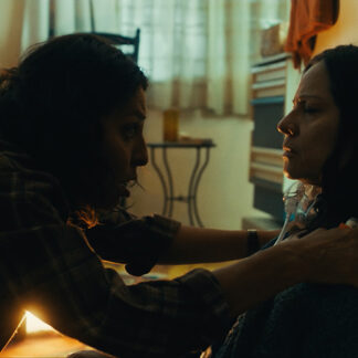 Two characters in a dimly lit room. One is leaning in urgently towards another individual who is seated against a wall. The seated person has dark hair and is wearing a thick knitted sweater, with a medical oxygen tube visible around their neck.