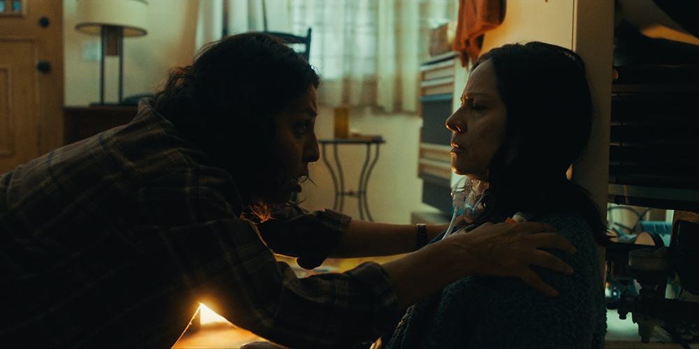 Two characters in a dimly lit room. One is leaning in urgently towards another individual who is seated against a wall. The seated person has dark hair and is wearing a thick knitted sweater, with a medical oxygen tube visible around their neck.