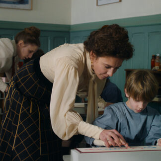 A still from "La Nouvelle Femme" focuses on a teacher guiding a young boy who is using a tactile learning aid, with another woman in the background. The room has a classic, early 20th-century aesthetic.