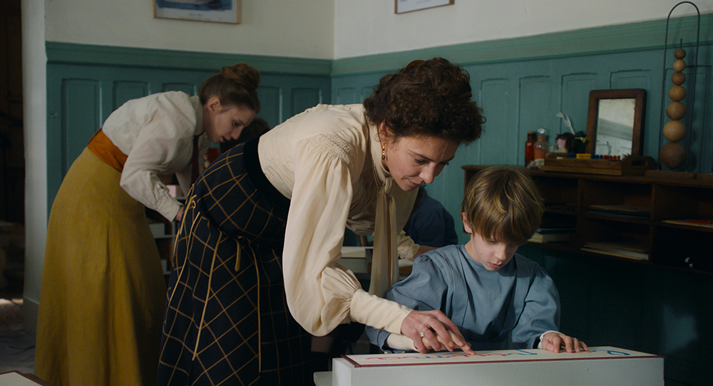 A still from "La Nouvelle Femme" focuses on a teacher guiding a young boy who is using a tactile learning aid, with another woman in the background. The room has a classic, early 20th-century aesthetic.