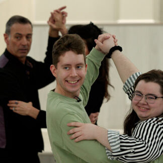 A still from "Lights-Camera-Friendship" showing four smiling individuals, including a man with a blue shirt and a young woman in glasses and a red jacket in the foreground, holding hands during a dance or activity.