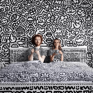 Mr doodle, a person with red hair, sits upright in bed next to his wife, a blonde person. They both wear doodle shirts, in doodled sheets, in a room with doodles all over the walls and furniture. The doodles are all in black and white