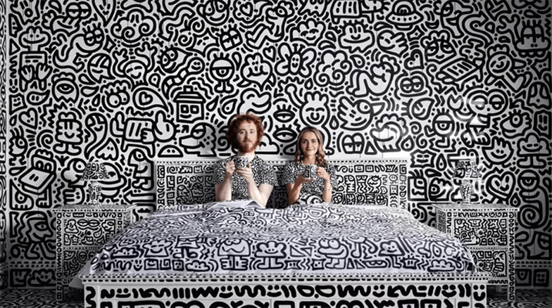 Mr doodle, a person with red hair, sits upright in bed next to his wife, a blonde person. They both wear doodle shirts, in doodled sheets, in a room with doodles all over the walls and furniture. The doodles are all in black and white
