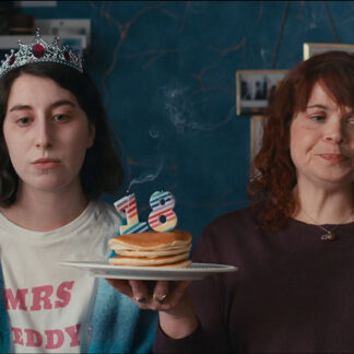 Two women side by side, one is wearing a tiara and a white shirt with pink text that reads "MRS TEDDY." The other is holding a plate with a stack of pancakes and two candles shaped like the number 18. They both have a grumpy expression.