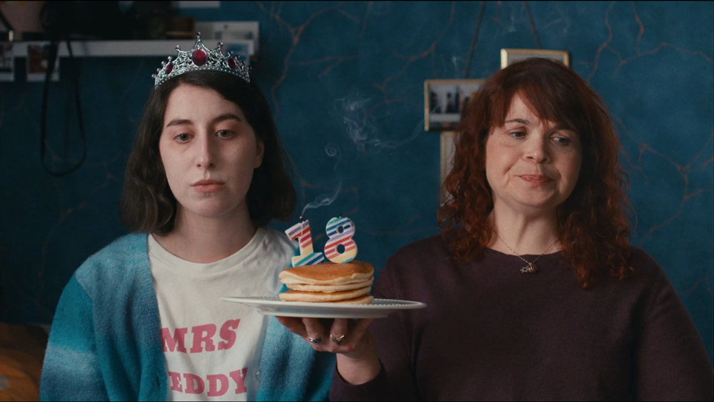 Two women side by side, one is wearing a tiara and a white shirt with pink text that reads "MRS TEDDY." The other is holding a plate with a stack of pancakes and two candles shaped like the number 18. They both have a grumpy expression.