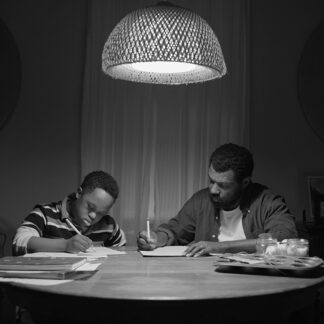 A black and white shot of a father and son working on homework together at a dimly lit table, illuminated by a woven light fixture overhead.