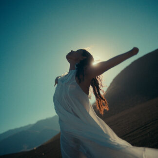 A woman with a limb difference, in a flowing white outfit standing outdoors with her arm outstretched, facing the sun with a serene expression.