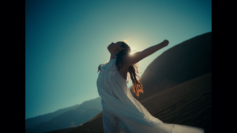 A woman with a limb difference, in a flowing white outfit standing outdoors with her arm outstretched, facing the sun with a serene expression.