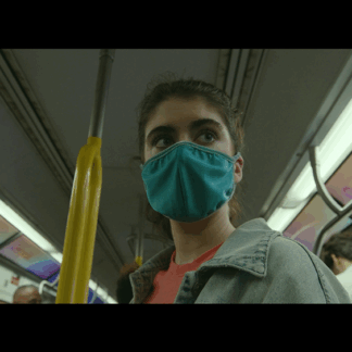 A young individual wearing a green medical mask that covers their mouth and nose, holding onto a pole in a subway car, gazing intensely at a distant point.