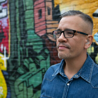 Person with glasses wearing a blue collard shirt standing in front of multi color graffiti wall
