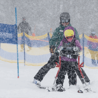 A child with adaptive ski equipment is assisted by an adult down a snow-covered ski slope. The child is wearing a yellow helmet and purple jacket. Snow is falling heavily. A banner for the Adaptive Sports Foundation is visible in the background.