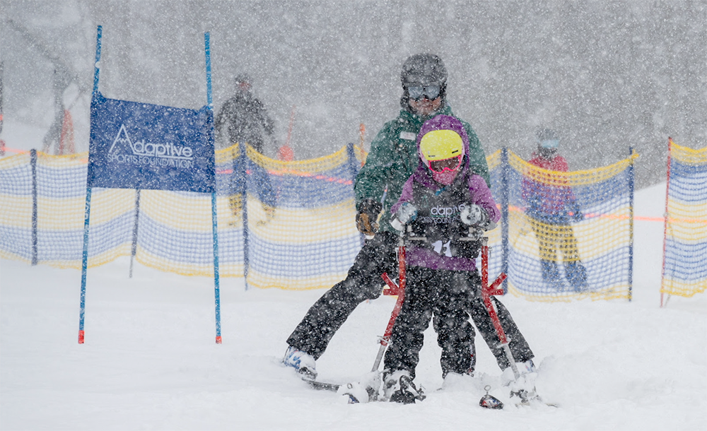 A child with adaptive ski equipment is assisted by an adult down a snow-covered ski slope. The child is wearing a yellow helmet and purple jacket. Snow is falling heavily. A banner for the Adaptive Sports Foundation is visible in the background.