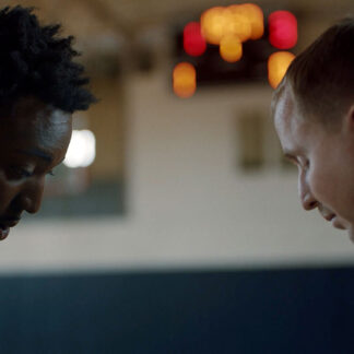 Profile view of a Black man with short curly hair and a light-skinned man with short hair, both wearing dark shirts. They are looking down, their faces close together, in an indoor setting with an out-of-focus scoreboard in the background.